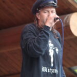 Male singer wearing a police-style hat performs on an outdoor wooden stage, holding a microphone.