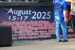 Event banner for August 15–17, 2025 with a long list of bands at the bottom and a person in a blue shirt standing to the right.