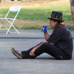 Man in a dark T-shirt and a black hat sits on the pavement outdoors, drinking from a blue can while nearby chairs and people stand in the background.